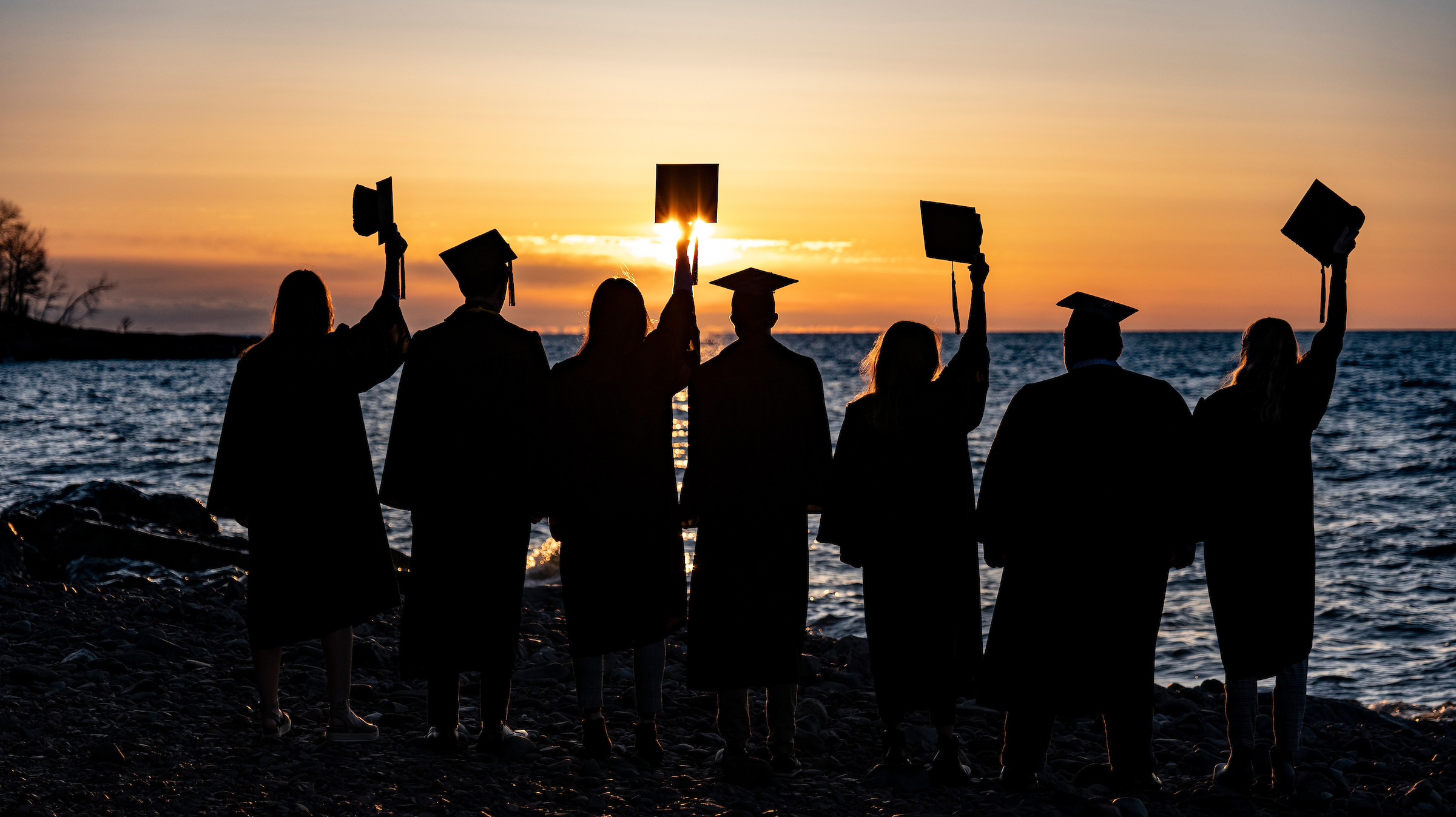 Seven students in cap and gown facing the sunrise over Lake Superior, with four waving their caps.